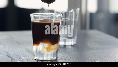 Gießen Sie Espresso in Tonic mit klarem Eiswürfel in Glas auf einem Tisch aus schwarzer Eiche Stockfoto