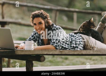 Eine Frau mit lockigen Haaren und einem karierten Hemd liegt auf einer Bank und arbeitet an einem Laptop. Ihre Katze liegt auf dem Rücken und eine Kaffeetasse ist neben ihr, c Stockfoto