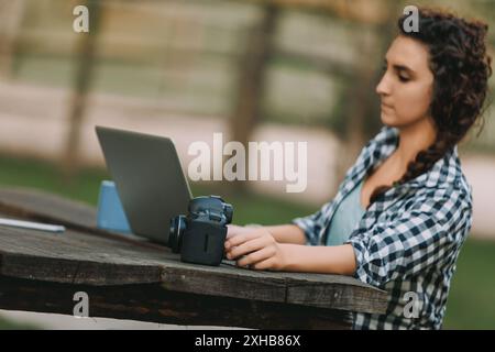 Eine Frau mit lockigen geflochtenen Haaren und einem karierten Hemd sitzt an einem Tisch im Freien und lächelt, während sie ihre Kamera untersucht. Ein Laptop ist vor ihr, Vorschlag Stockfoto