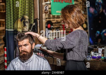 Friseurmädchen rasiert seinen Bart mit einem Haarschneider. Friseur für Frauen. Friseurfrau schneidet einen bärtigen Mann in einem Friseurladen. Mann besucht Stockfoto