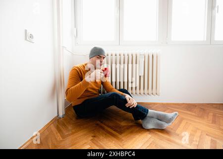 Ein Mann in einer Mütze sitzt auf dem Boden neben einem Heizkörper und trinkt aus einem roten Becher. Stockfoto