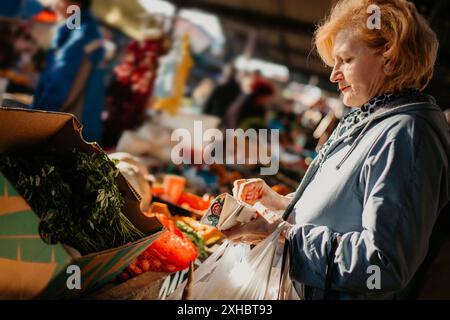Eine Frau inspiziert sorgfältig eine Schachtel frisches grünes Gemüse, während sie auf einem Markt im Freien einkaufen. Stockfoto