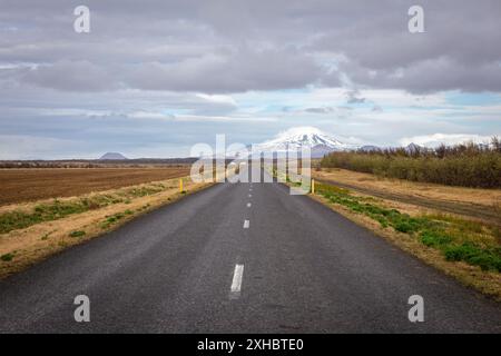 Gerade Asphaltstraße durch das Grasland des Südens Islands mit gelben Straßenmarkierungen und schneebedecktem Hekla-Vulkan im Hintergrund. Stockfoto