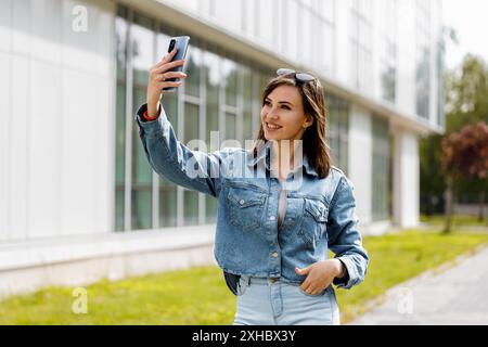 Eine Frau macht ein Selfie mit ihrem Smartphone, während sie an einem sonnigen Tag vor einem modernen Gebäude läuft. Stockfoto