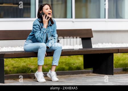 Eine Frau sitzt auf einer Bank vor einem Gebäude, spricht mit ihrem Handy und lacht. Stockfoto