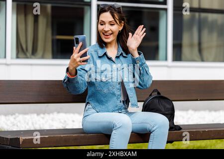 Eine junge Frau sitzt auf einer Bank und benutzt ihr Smartphone, um jemanden per Video anzurufen, und winkt mit ihrer rechten Hand. Stockfoto