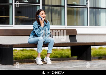 Eine junge Frau mit Jeansjacke und blauer Jeans sitzt auf einer Bank vor einem modernen Gebäude, während sie mit einem Smartphone spricht. Stockfoto
