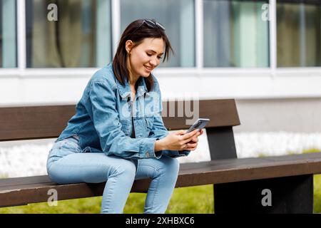 Eine junge Frau in Jeans und Jeansjacke sitzt auf einer Bank und lächelt, während sie ihr Smartphone benutzt. Stockfoto