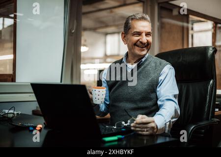 Ein Mann sitzt an seinem Schreibtisch in einem Büro und lächelt, während er nach rechts blickt. Er hält einen Becher in der Hand und schaut sich etwas aus der Kamera an. Stockfoto
