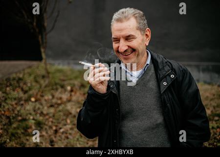 Ein Mann mittleren Alters mit grauen Haaren, trägt eine schwarze Jacke und einen grauen Pullover, lächelt, während er eine Zigarette hält. Stockfoto