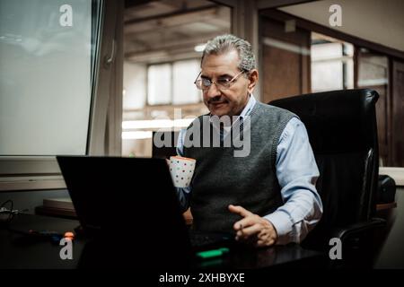 Ein Mann sitzt in einem schwarzen Bürostuhl und schaut auf seinen Laptop, mit einer Kaffeetasse in der rechten Hand. Er trägt ein blaues Hemd und eine graue Weste. Stockfoto