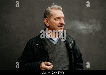 Ein Mann mittleren Alters mit grauen Haaren in einer schwarzen Jacke raucht eine Zigarette, während er vor einer grauen Wand steht. Stockfoto