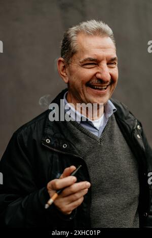 Ein lächelnder älterer Mann mit Schnurrbart hält eine Zigarette in der rechten Hand. Stockfoto
