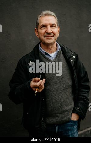 Ein älterer Mann in einer schwarzen Jacke und einem grauen Pullover raucht eine Zigarette, während er nach oben blickt. Stockfoto
