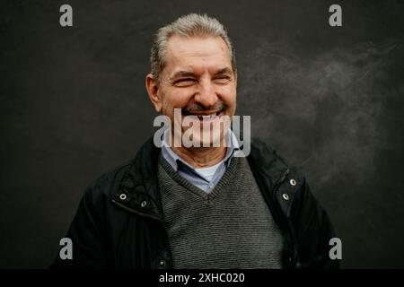 Ein Mann mit grauen Haaren und Schnurrbart lächelt breit, während er eine schwarze Jacke über einem grauen Pullover trägt. Stockfoto