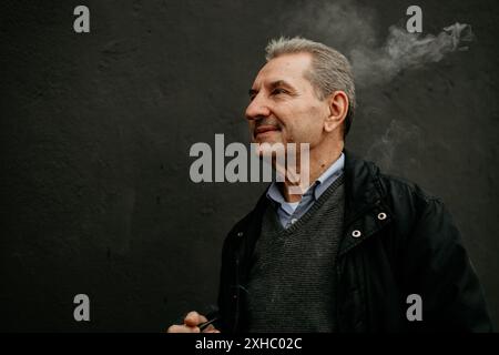 Ein Mann in einer schwarzen Jacke und einem grauen Pullover raucht eine Pfeife mit Rauch, der sich über seinem Kopf kräuselt. Stockfoto