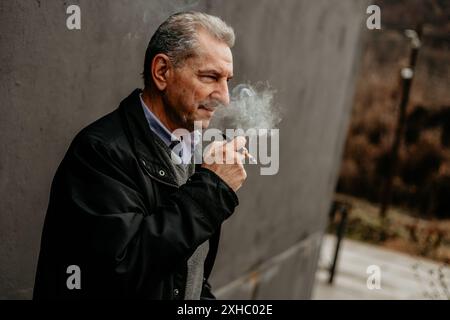 Ein Mann in einer schwarzen Jacke raucht eine Zigarette, während er an einer grauen Wand steht. Stockfoto