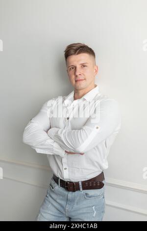 Hübscher erfolgreicher junger Mann mit Frisur in einem modischen weißen Hemd mit Jeans steht neben einer weißen Wand Stockfoto