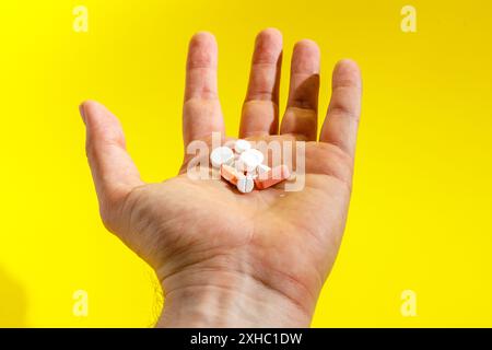 Eine Hand hält einen kleinen Stapel Pillen, darunter weiße runde Pillen und rosafarbene rechteckige Pillen, auf gelbem Hintergrund. Stockfoto