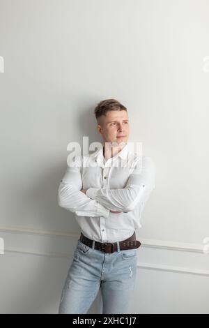 Ein erfolgreicher, gutaussehender junger Mann mit einer Frisur in einem modischen weißen Hemd mit Jeans steht an einer weißen Wand Stockfoto