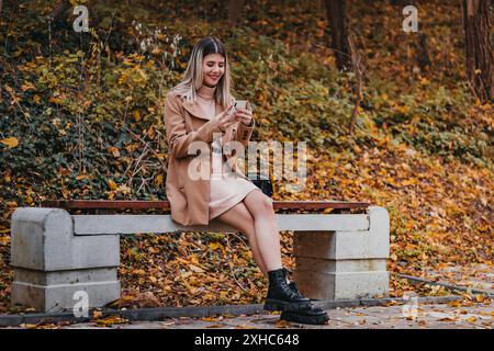 Eine Frau im braunen Mantel sitzt auf einer Bank im Park und benutzt ihr Telefon. Sie trägt ein pinkfarbenes Kleid und schwarze Stiefel. Der Boden ist mit abgefallenem Laub bedeckt Stockfoto