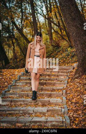 Eine Frau geht Steintreppen in einem Wald hinunter, trägt einen braunen Mantel und schwarze Stiefel. Der Boden ist mit Herbstlaub bedeckt. Stockfoto