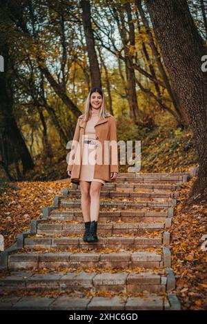 Eine Frau in braunem Mantel geht Steintreppen hinunter und lächelt, während sie nach vorne blickt. Gefallene Blätter liegen auf dem Boden und Stufen. Stockfoto