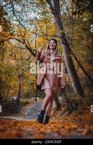 Eine Frau in braunem Mantel geht durch einen Herbstwald und lächelt, während sie ein gefallenes Blatt hält. Stockfoto