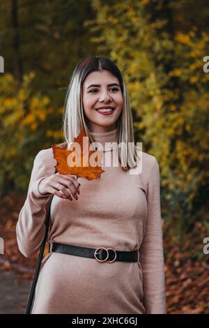 Eine Frau lächelt, als sie ein herabfallendes Herbstblatt hält und während der Herbstsaison in einem Park steht. Stockfoto