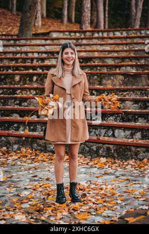 Eine Frau im braunen Mantel lächelt, während sie im Park steht und mit Herbstlaub spielt. Stockfoto