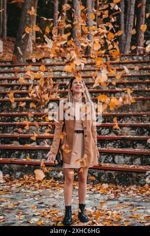 Eine Frau in braunem Mantel und Kleid steht auf Steintreppen und lächelt, während Blätter um sie herum fallen. Stockfoto