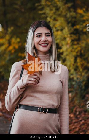 Eine Frau lächelt, als sie ein großes Orangenblatt in einem Park hält. Sie trägt ein langärmeliges, hellrosa Kleid mit einem schwarzen Gürtel. Stockfoto