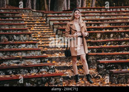 Eine Frau in einem braunen Mantel steht auf einer Steintreppe, die mit Herbstlaub bedeckt ist, und lächelt in die Kamera. Stockfoto