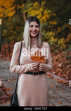 Eine Frau in einem rosafarbenen Kleid mit langen Ärmeln lächelt, während sie ein braunes Blatt im Park hält. Stockfoto
