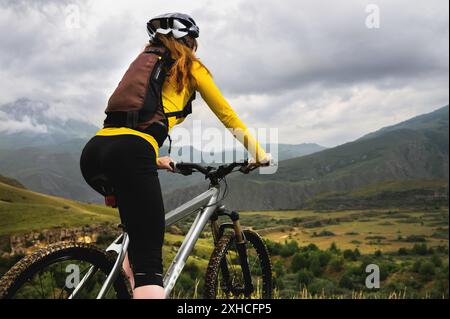 Ein junges schlankes Mädchen mit Rucksack und Fahrradhelm sitzt an einem bewölkten Tag auf einem Fahrrad hoch in den Bergen und schaut auf die Berge und Stockfoto