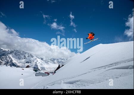 Ein junger männlicher Profisportler in einem orangen Skianzug fliegt über die Berge, nachdem er von schneebedeckten Felsvorsprüngen gesprungen ist. Freeridesport Stockfoto