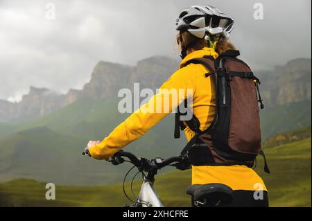 Nahaufnahme eines jungen schlanken Mädchens mit Rucksack und Fahrradhelm sitzt an einem bewölkten Tag auf einem Fahrrad hoch in den Bergen und schaut auf die Stockfoto