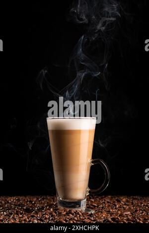 Heiß dampfender Kaffee Latte mit schaumiger Milch in hohem Glas, auf Kaffeebohnen auf schwarzem Hintergrund. Blick von oben Stockfoto
