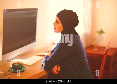Seitenansicht des nachdenklichen jungen Weibchens in schwarzer Kopfbedeckung, das vor dem großen Computermonitor am Tisch mit einer Tasse Kaffee sitzt, während er zu Hause arbeitet Stockfoto