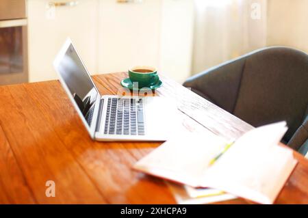 Moderner Laptop und offene Notizblöcke auf Küchentisch in der Nähe einer Tasse Kaffee zu Hause platziert Stockfoto