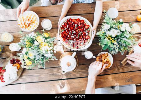 Blick von oben auf eine nicht erkennbare Familie, die am Holztisch mit Waffeln und Erdbeeren saß und am sonnigen Morgen ein köstliches Frühstück hatte Stockfoto