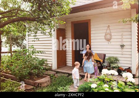 Junge, entzückte Mutter sitzt im Sessel mit entzückenden kleinen Geschwistern auf der Terrasse des Landhauses und entspannt sich am Wochenende zusammen Stockfoto