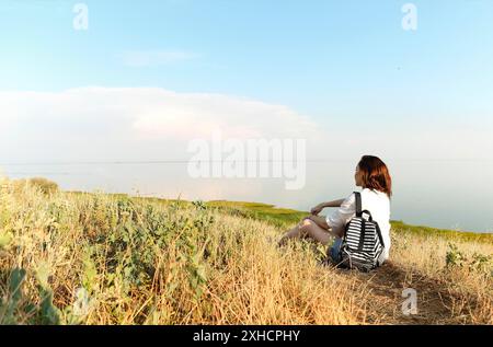 Seitenansicht der unkenntlichen weiblichen Reisenden auf Gras auf dem Hügel sitzen und bewundern erstaunliche Landschaft während Sonnenuntergang im Sommer Stockfoto