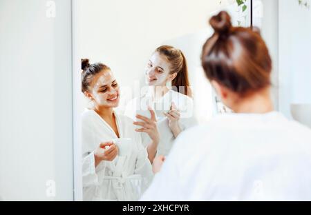 Lächelnde Freundinnen in Bademänteln und mit Lehmmasken im Gesicht im Badezimmer stehend und im Spiegel auf dem Smartphone fotografieren Stockfoto