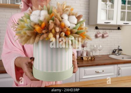 Eine junge Frau in einem rosafarbenen Bademantel, die eine runde Geschenkbox mit getrockneten Blumen in der Küche hält. Die hellgrüne Box mit weißen Streifen befindet sich auf einer Holzlasche Stockfoto