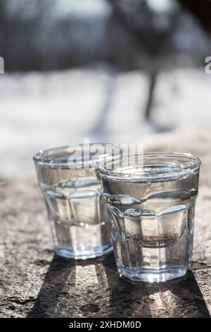 Zwei Klarglas-Shot-Gläser, gefüllt mit Wasser auf Einer Holzoberfläche im Sonnenlicht Stockfoto