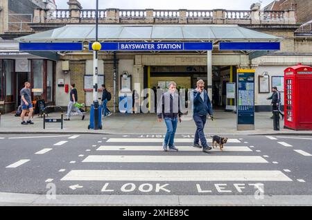 Männer, die einen Hund laufen, überqueren die Straße über einen Fußgängerübergang vor der Bayswater London U-Bahnstation am Queensway Stockfoto