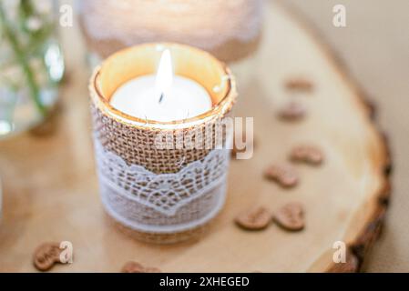 Warm beleuchtete Kerze in brauner Leinwand und weißer Spitze auf Baumrinde und Miniaturherzen im Hintergrund Stockfoto