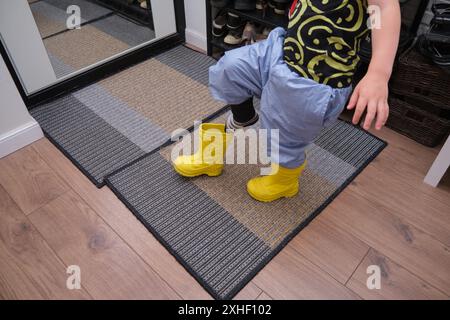 Ein Kind zieht Schuhe an, die im Flur stehen. Das Baby zieht gelbe Stiefel an, um spazieren zu gehen. Kind im Alter von 3 Jahren (dreijähriger Junge) Stockfoto