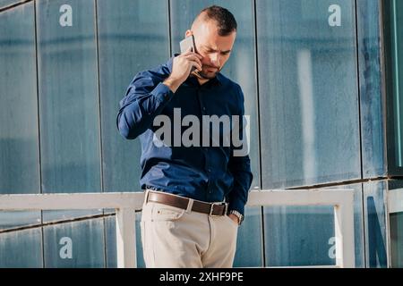 Ein Mann in einem blauen Button-Down-Hemd und einer beigefarbenen Hose spricht vor einem modernen Gebäude auf seinem Handy. Stockfoto